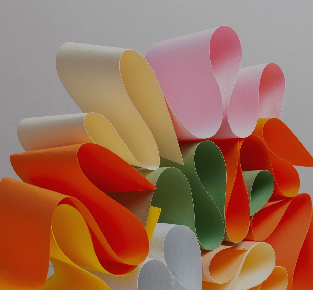 A cluster of curled paper sheets in many different colours, pink, yellow, orange, green, and cream, fanned together against a neutral grey background.