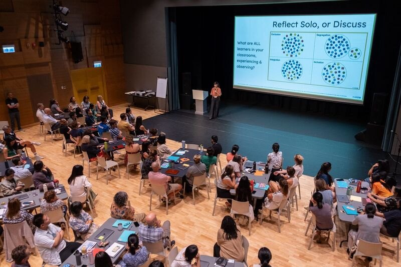 Elevated view of Beth Stark presenting to a large seated audience in a tiered auditorium; the screen displays "Reflect Solo, or Discuss" with circular diagrams. Image courtesy of Hong Kong Academy.